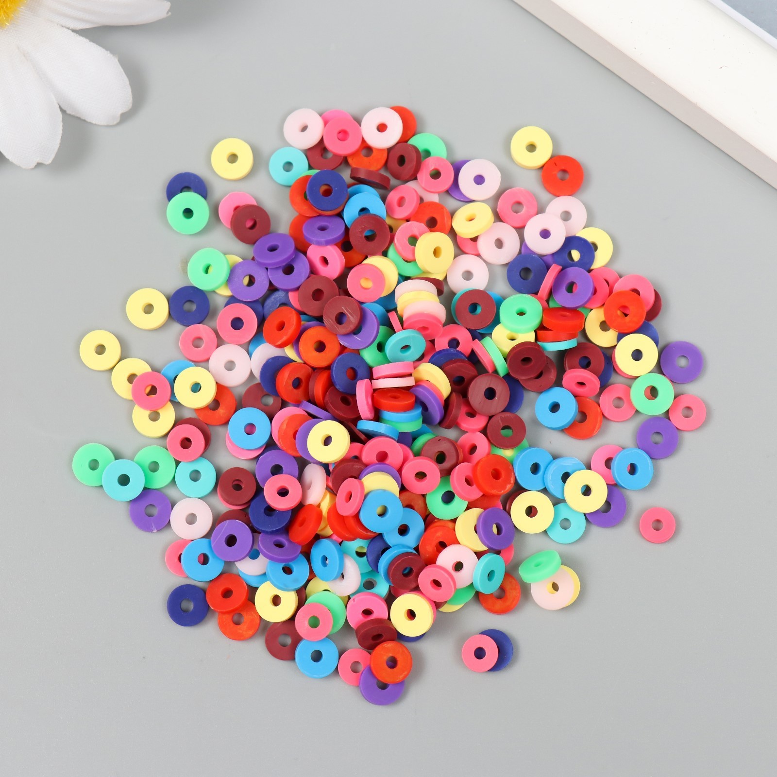 Россыпь мелких плоских круглых бусин. Они разноцветные — есть розовые, фиолетовые, синие, красные, белые, оранжевые и другие оттенки. У каждой бусины в центре есть отверстие. Фон — светло-серый.