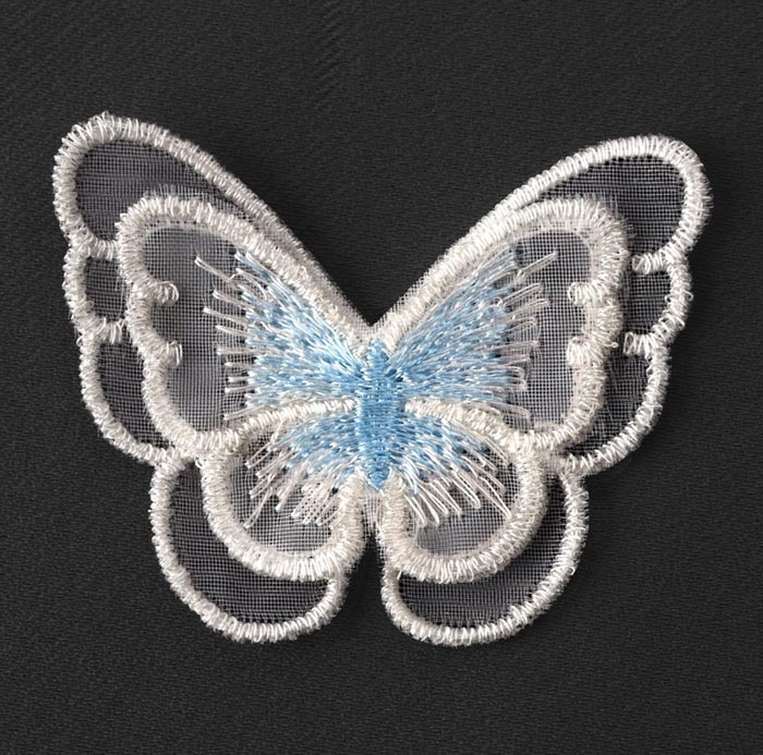 На тёмно-сером фоне лежит нашивка в форме бабочки. Она выполнена на полупрозрачной сетчатой основе. Контуры крыльев вышиты белыми нитками, а тельце — тёмно-зелёными или чёрными, с добавлением прозрачных блестящих элементов, похожих на леску.